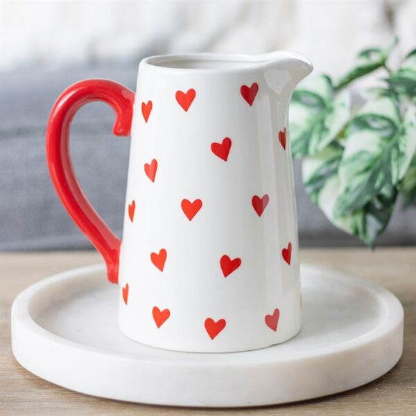 A charming white pitcher with a red handle and scattered red heart patterns, resting on a tray.