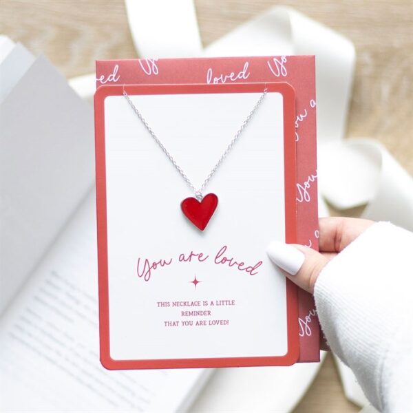 A hand holds a card featuring a heart-shaped necklace with the message “You are loved.”