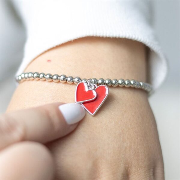 A close-up of a hand touching a silver bracelet with two red heart charms.