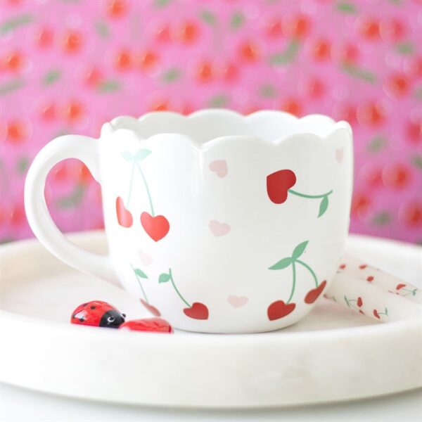 A cute scalloped cup decorated with cherries sits on a white plate beside a ladybug.