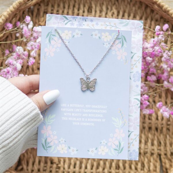 A hand holds a butterfly necklace on a floral greeting card with inspirational text.